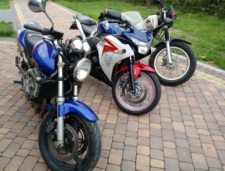 Trzy motocykle różnych marek i modeli zaparkowane obok siebie na kostce brukowej: niebieski naked bike z przodu, biało-czerwony sportowy motocykl pośrodku i czarny motocykl enduro z prawej strony.