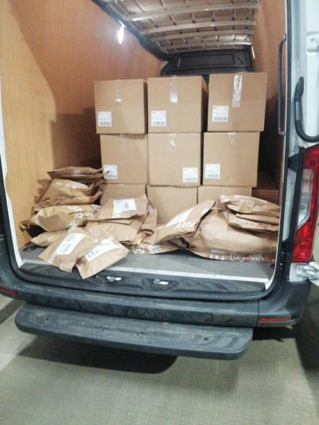 Wnętrze furgonetki wypełnionej kartonowymi pudłami i brązowymi paczkami, widok z tyłu pojazdu.