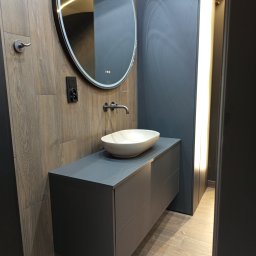 Meblebolo Przemysław Fuczyło - Nowoczesna łazienka z szarą szafką pod umywalkę, owalnym lustrem z podświetleniem LED i drewnopodobnymi płytkami na ścianie. Minimalistyczny design.