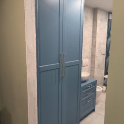 Meblebolo Przemysław Fuczyło - Niebieska szafa łazienkowa z złotymi uchwytami, umywalką nablatową i widoczną instalacją sanitarną w tle. Świeżo wykończone obramowanie drzwi.