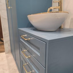 Meblebolo Przemysław Fuczyło - Niebieska szafka łazienkowa z trzema szufladami i złotymi uchwytami, na której stoi biała umywalka ze złotym obramowaniem i złotą baterią. Widoczne fragmenty ściany i podłogi.