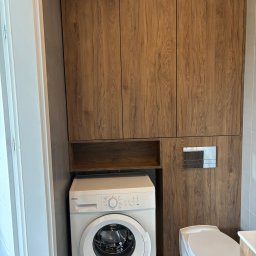 Meblebolo Przemysław Fuczyło - Drewniana zabudowa w łazience z szafkami nad pralką i toaletą. Fronty z wyraźnym rysunkiem drewna, szara ściana i biała pralka Amica. Minimalistyczny styl.