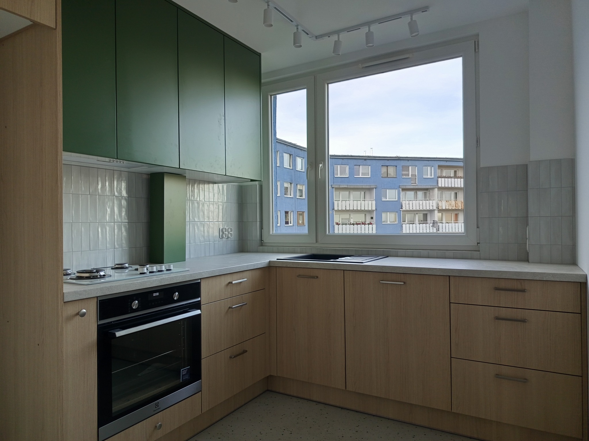 Jasna kuchnia z drewnianymi szafkami i zielonymi górnymi szafkami. Widok na blok z okna. Minimalistyczny design, białe płytki na ścianie.