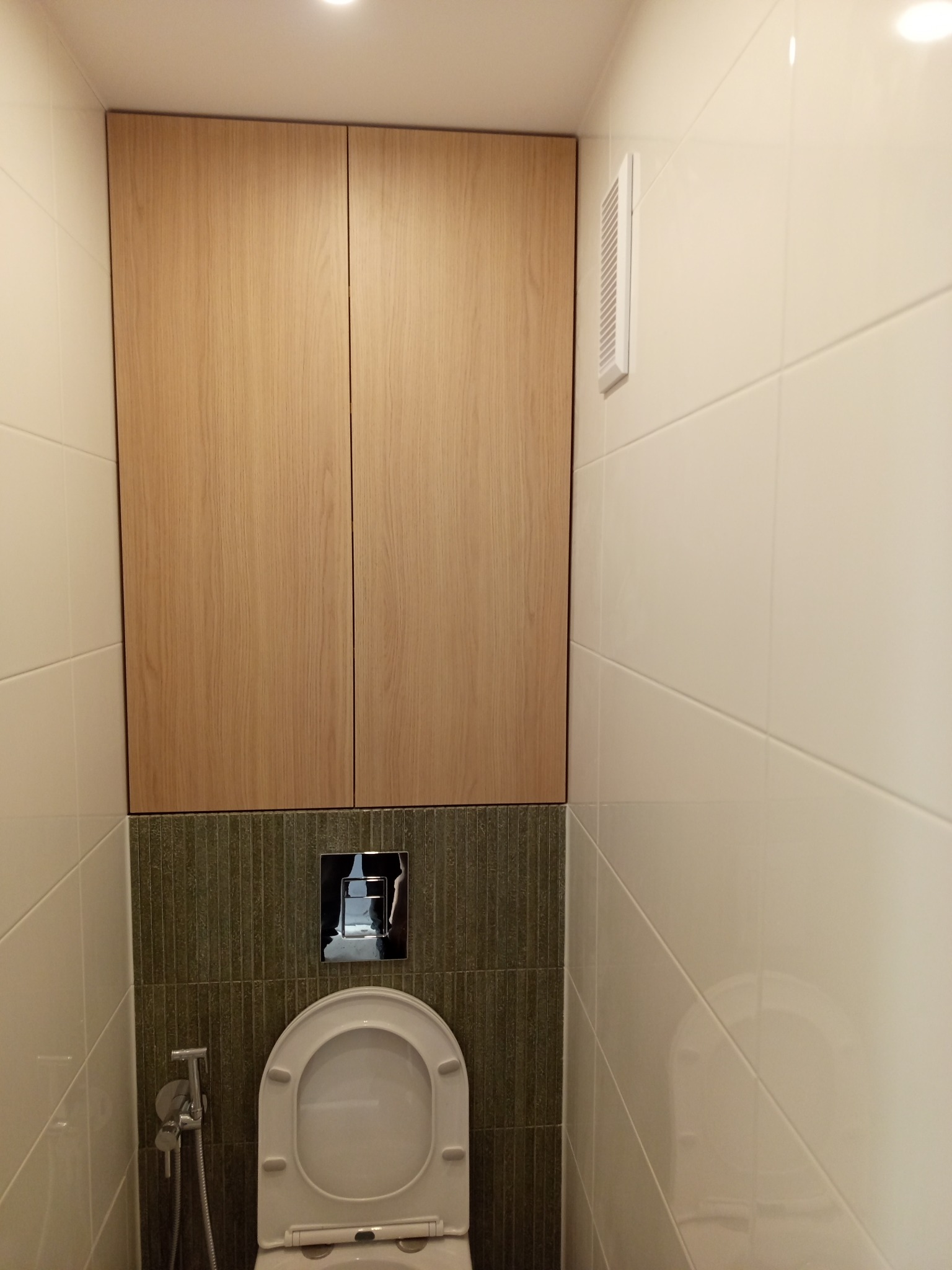 Szafka łazienkowa z drewnianymi frontami nad toaletą, wykończona płytkami w odcieniach bieli i zieleni. Widoczny chromowany przycisk spłuczki i bidetka.