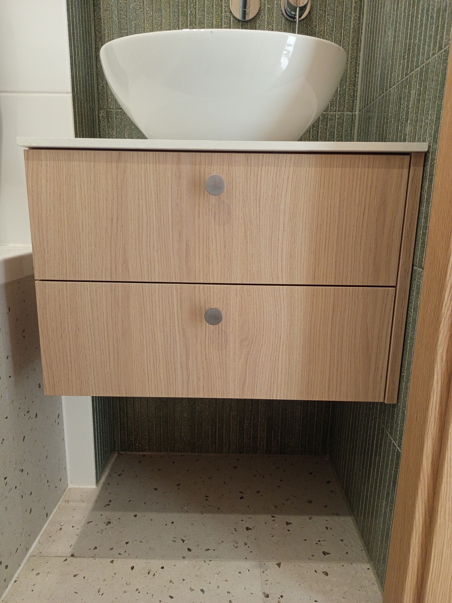 Nowoczesna szafka łazienkowa z dwoma szufladami o drewnianym wykończeniu, umywalką nablatową i zielonymi płytkami w tle. Minimalistyczny design.