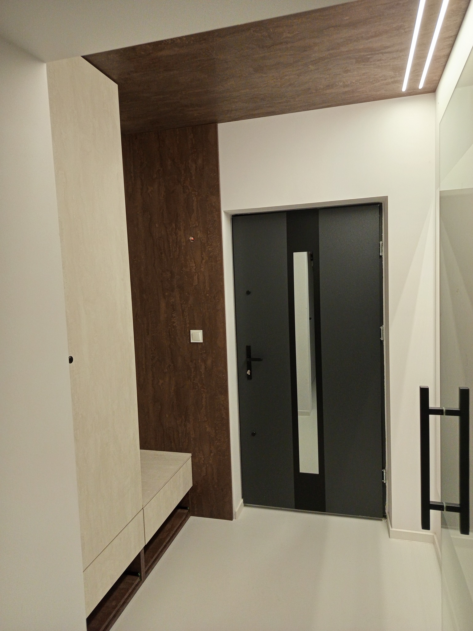 Nowoczesna szafa wnękowa z siedziskiem w przedpokoju, wykończona w beżowych i brązowych odcieniach, z minimalistycznym oświetleniem sufitowym i ciemnymi drzwiami wejściowymi.