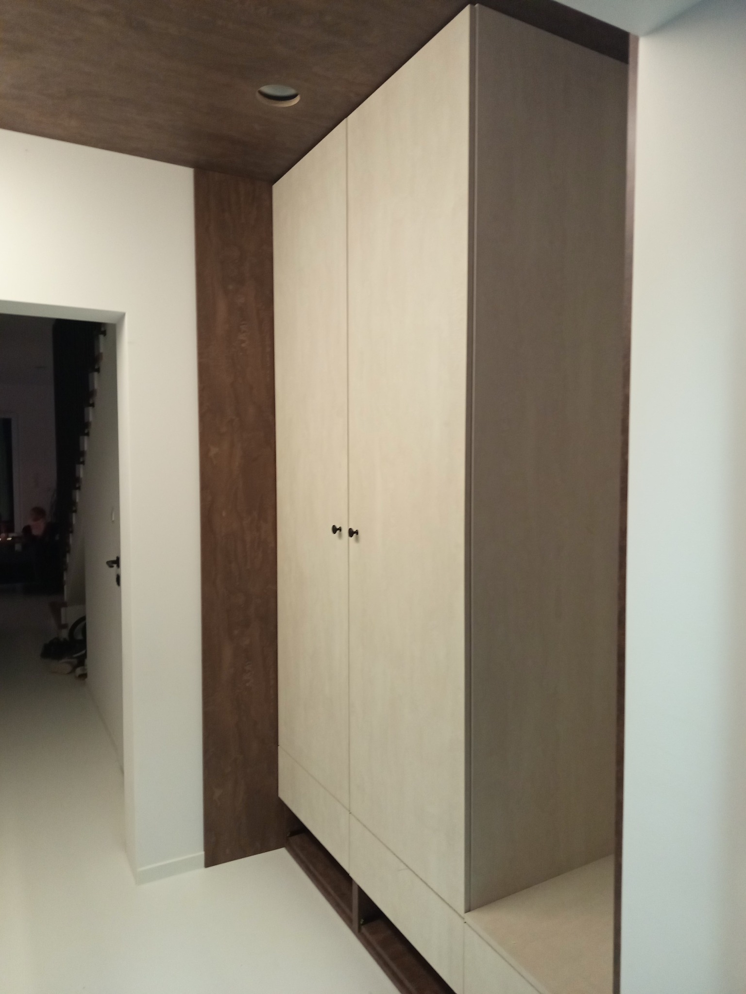 Dwudrzwiowa szafa wnękowa z jasnymi frontami i ciemnobrązowymi bokami, wkomponowana w korytarz z drewnianym sufitem i białymi ścianami. Minimalistyczny design.