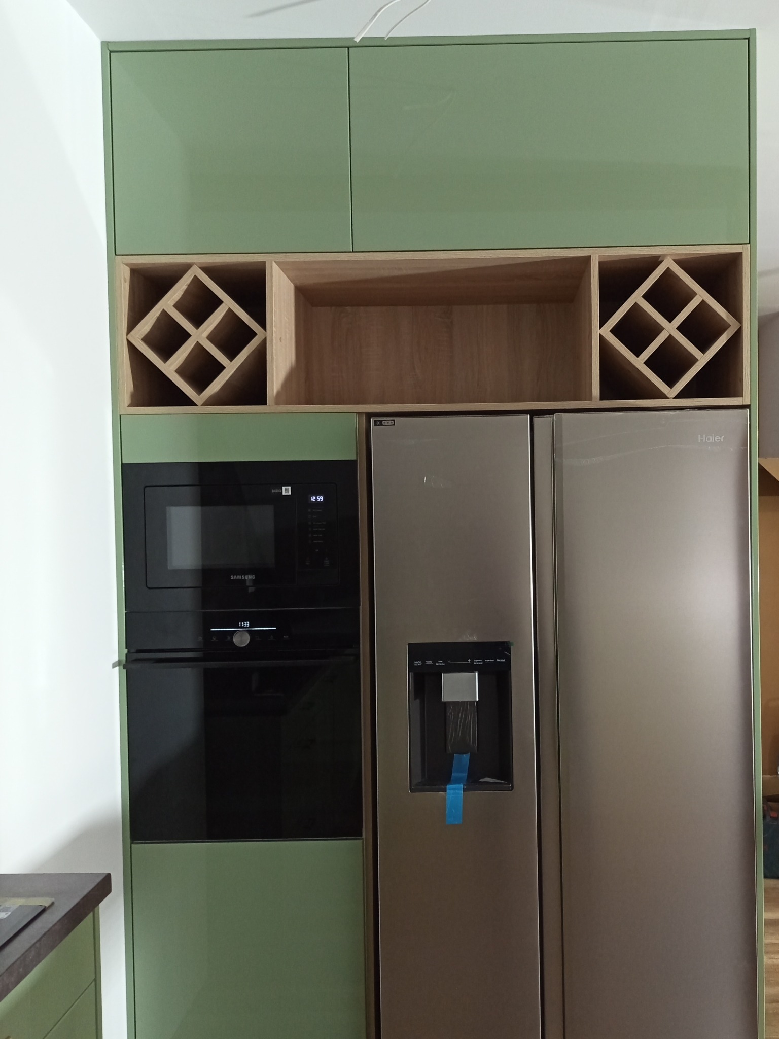 Nowoczesna kuchnia na wymiar: pistacjowe szafki, drewniane półki na wino, zabudowana lodówka side-by-side i piekarnik z mikrofalówką. Minimalistyczny design.