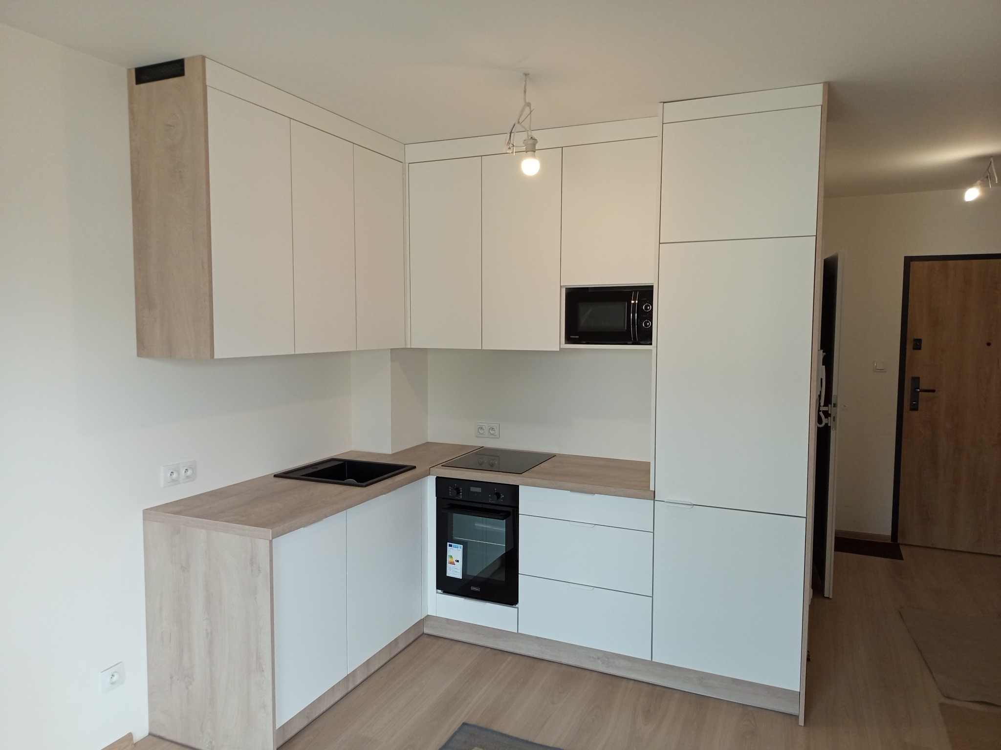 Nowoczesna, narożna kuchnia na wymiar z białymi frontami i blatem w odcieniu jasnego drewna. Widoczny piekarnik, płyta indukcyjna i mikrofalówka w zabudowie.