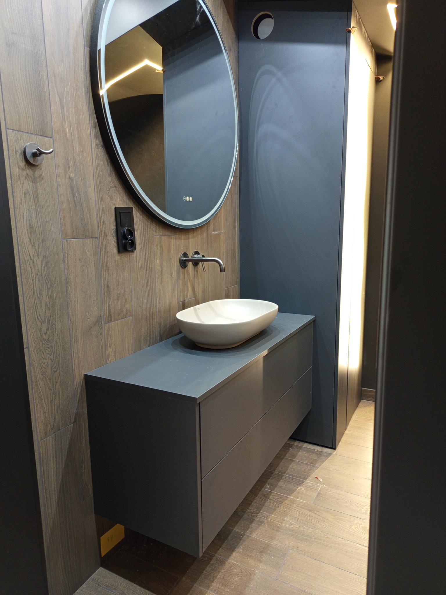 Nowoczesna łazienka z szarą szafką pod umywalkę, owalnym lustrem z podświetleniem LED i drewnopodobnymi płytkami na ścianie. Minimalistyczny design.