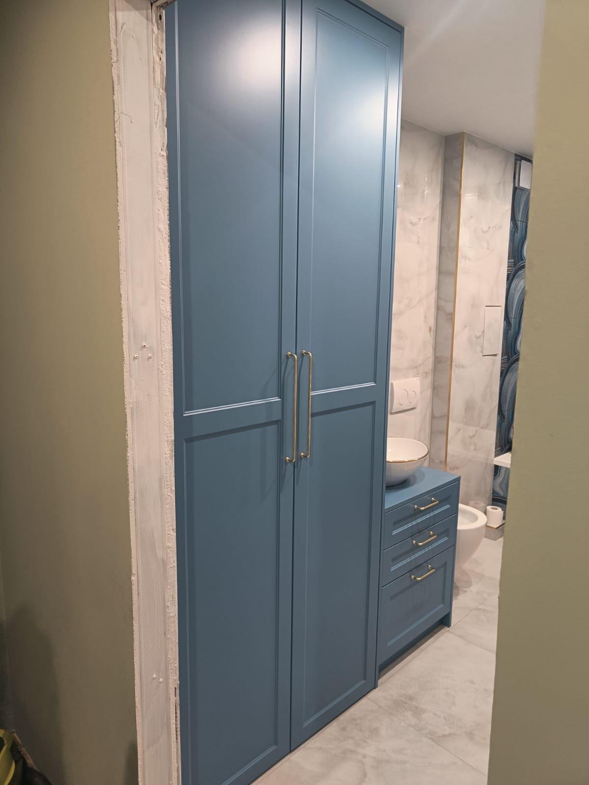 Niebieska szafa łazienkowa z złotymi uchwytami, umywalką nablatową i widoczną instalacją sanitarną w tle. Świeżo wykończone obramowanie drzwi.