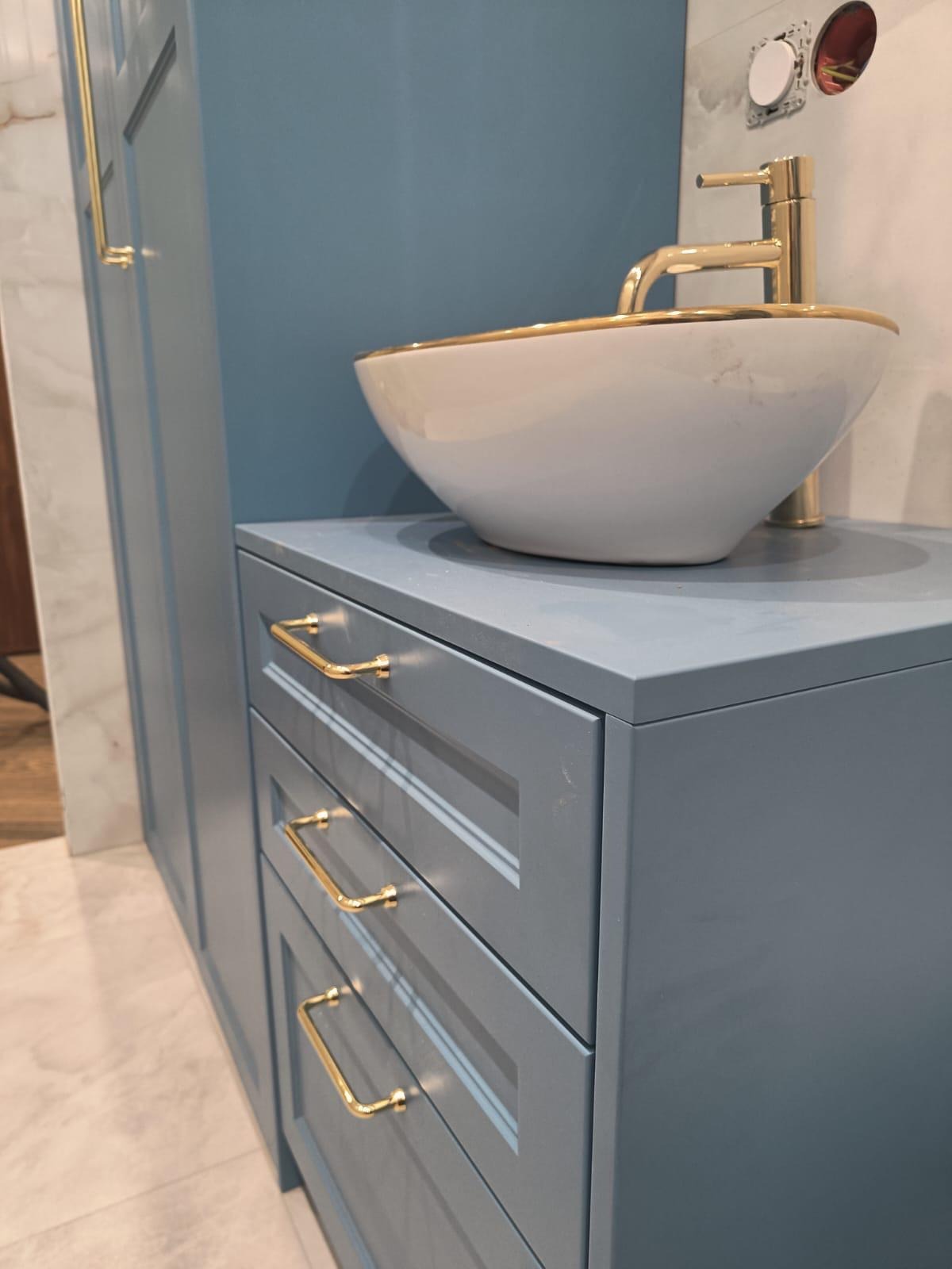 Niebieska szafka łazienkowa z trzema szufladami i złotymi uchwytami, na której stoi biała umywalka ze złotym obramowaniem i złotą baterią. Widoczne fragmenty ściany i podłogi.