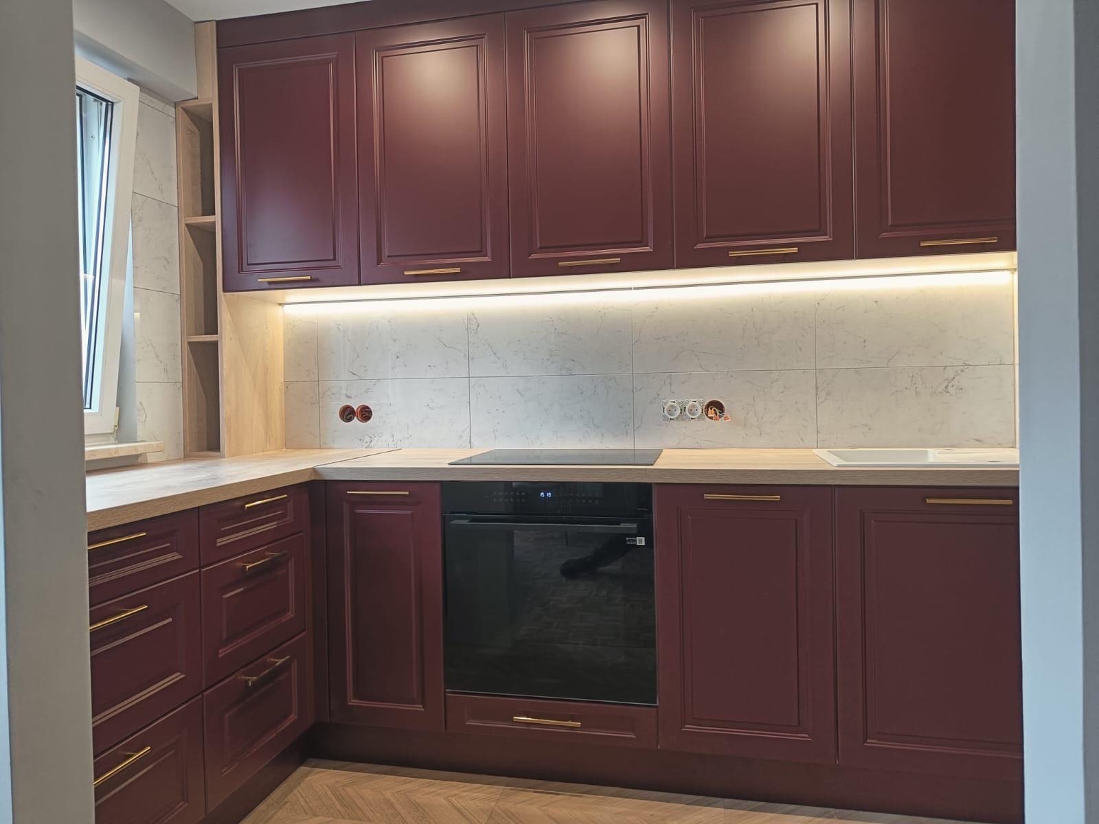 Bordowa kuchnia z frezowanymi frontami, złotymi uchwytami i blatem imitującym drewno. Nad blatem marmurowe płytki i oświetlenie LED. Widoczne gniazdka elektryczne.
