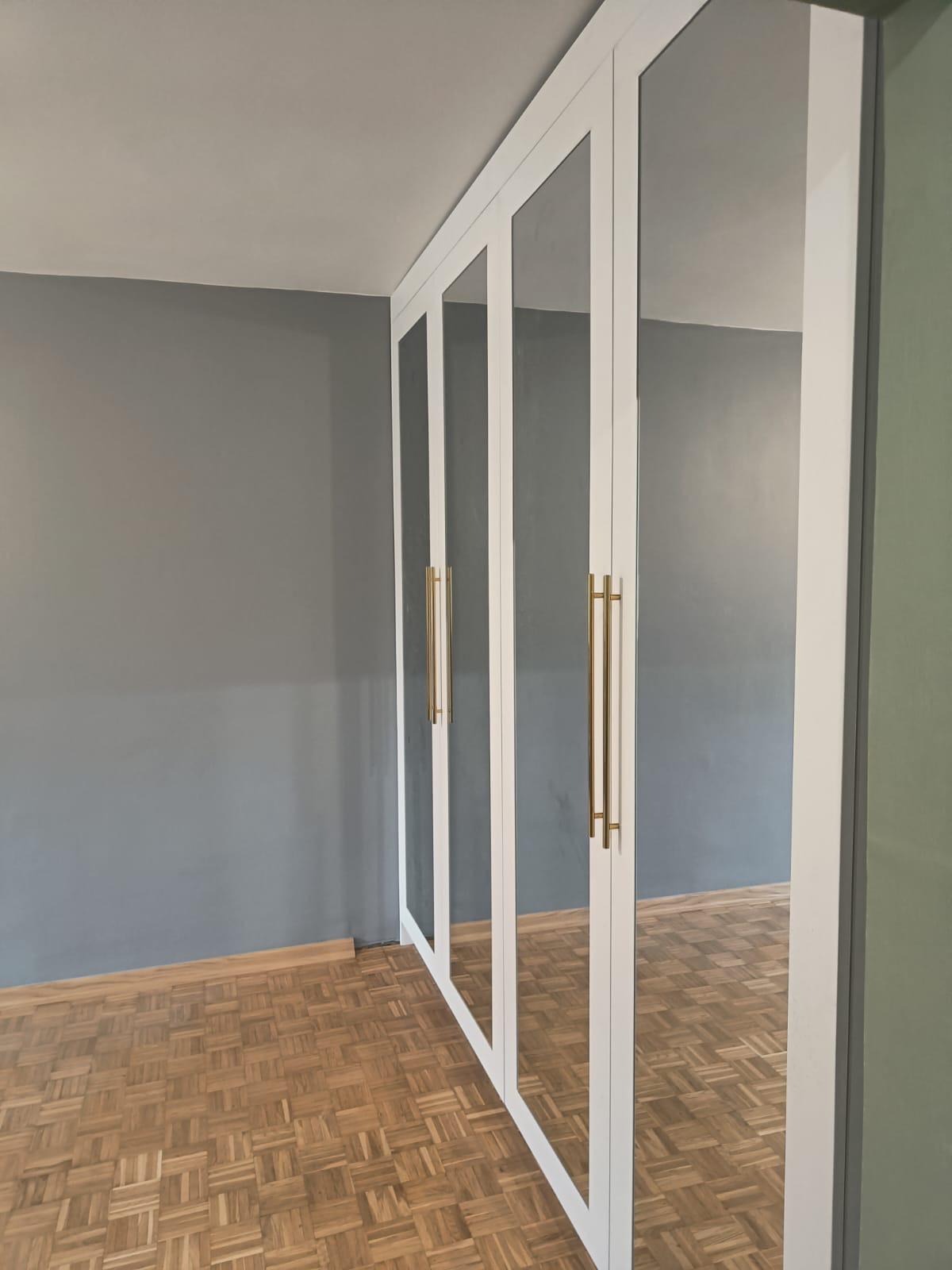 Biała szafa wnękowa z lustrzanymi drzwiami i złotymi uchwytami, parkiet na podłodze, szara ściana. Minimalistyczny design, nowoczesne wykończenie.