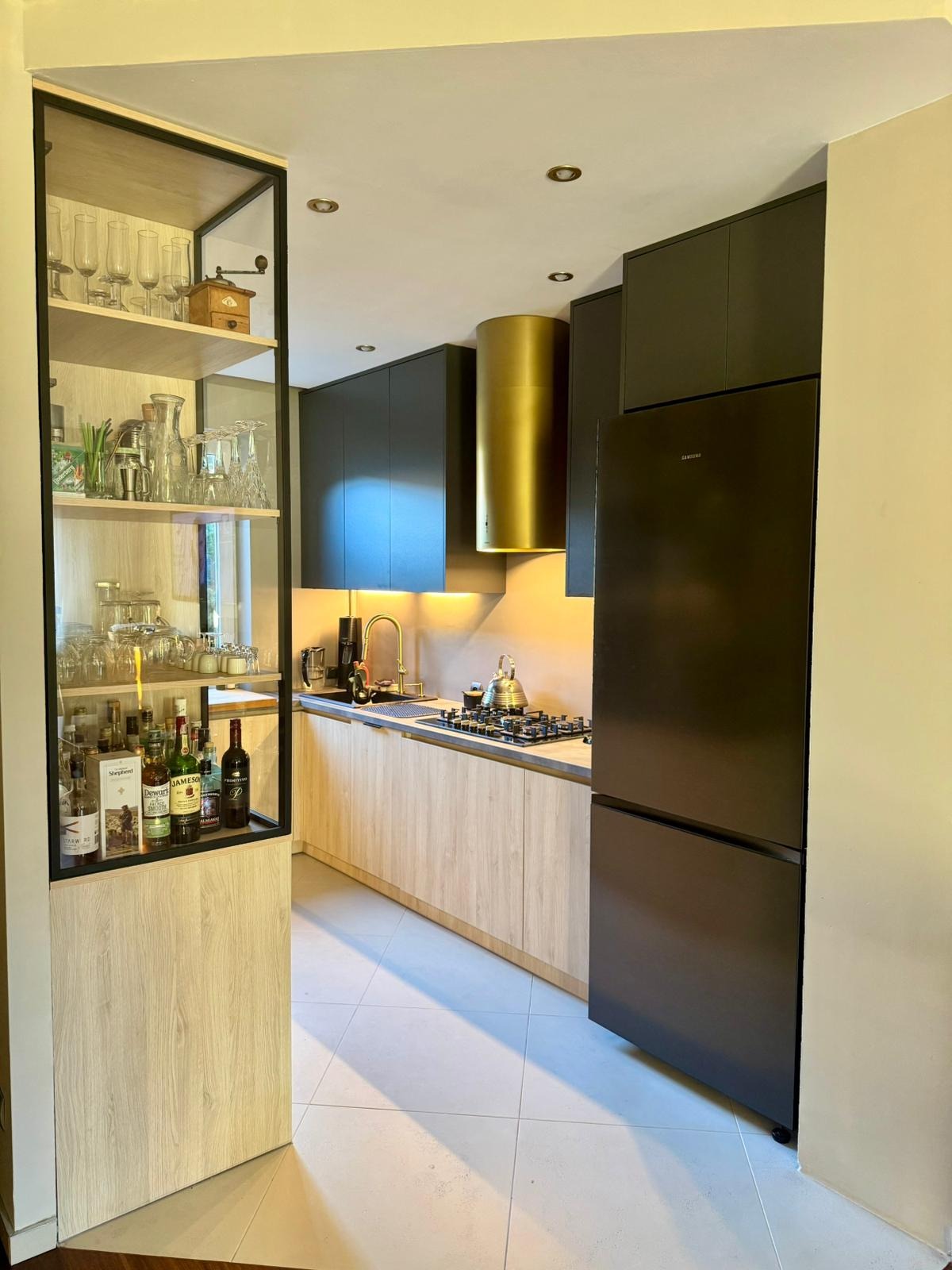 Nowoczesna kuchnia z szarymi szafkami górnymi i drewnopodobnymi dolnymi, złoty okap cylindryczny, przeszklona witryna z alkoholem i szkłem, czarna lodówka.