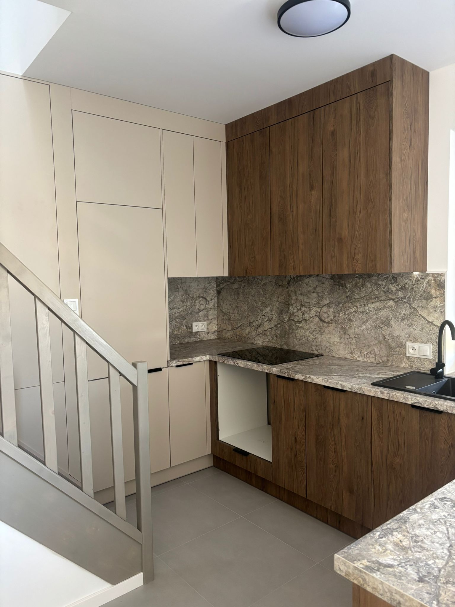 Dwukolorowa zabudowa kuchenna z drewnianymi szafkami górnymi i beżowymi dolnymi, marmurowy blat i ściana nad blatem, widok z perspektywy schodów.