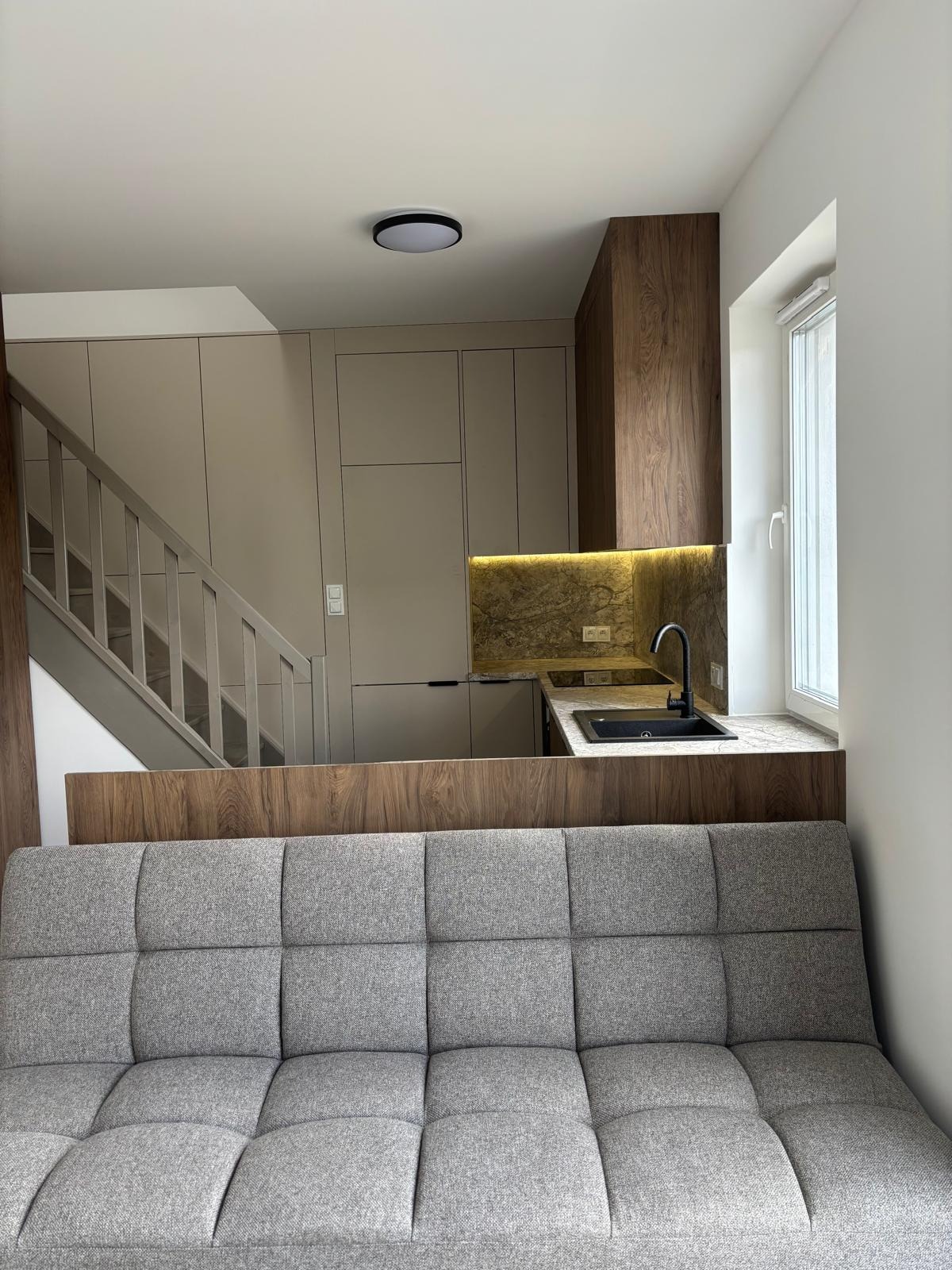 Nowoczesna kuchnia z beżowymi szafkami i drewnianym blatem, widoczna zza szarej sofy. Uwagę przykuwa oświetlenie LED pod szafkami i czarna bateria.