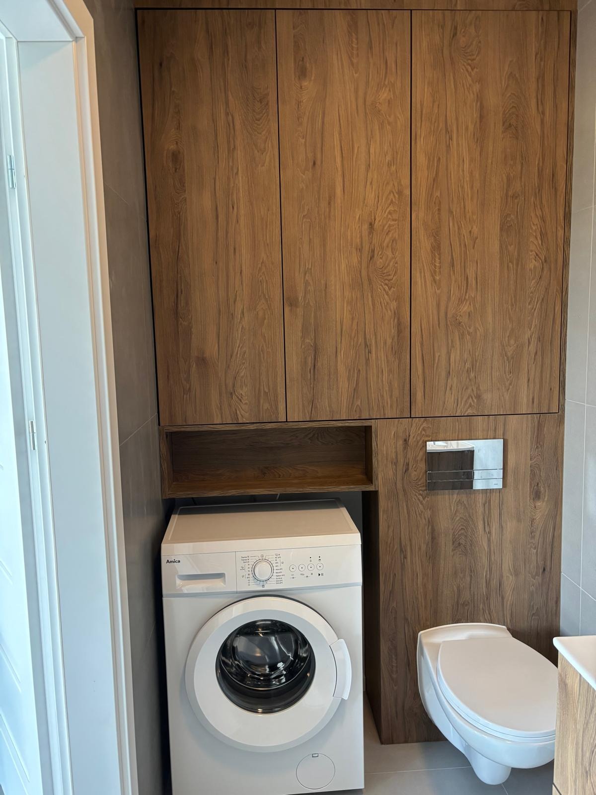 Drewniana zabudowa w łazience z szafkami nad pralką i toaletą. Fronty z wyraźnym rysunkiem drewna, szara ściana i biała pralka Amica. Minimalistyczny styl.