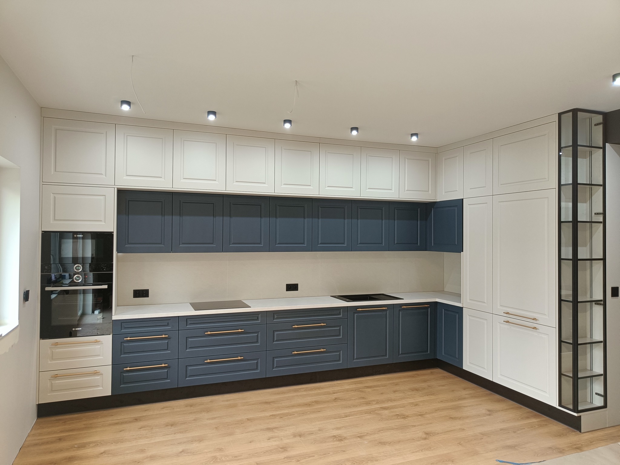 Nowoczesna kuchnia z białymi górnymi szafkami i granatowymi dolnymi, złotymi uchwytami. Widoczny piekarnik i płyta indukcyjna. Podłoga drewniana.