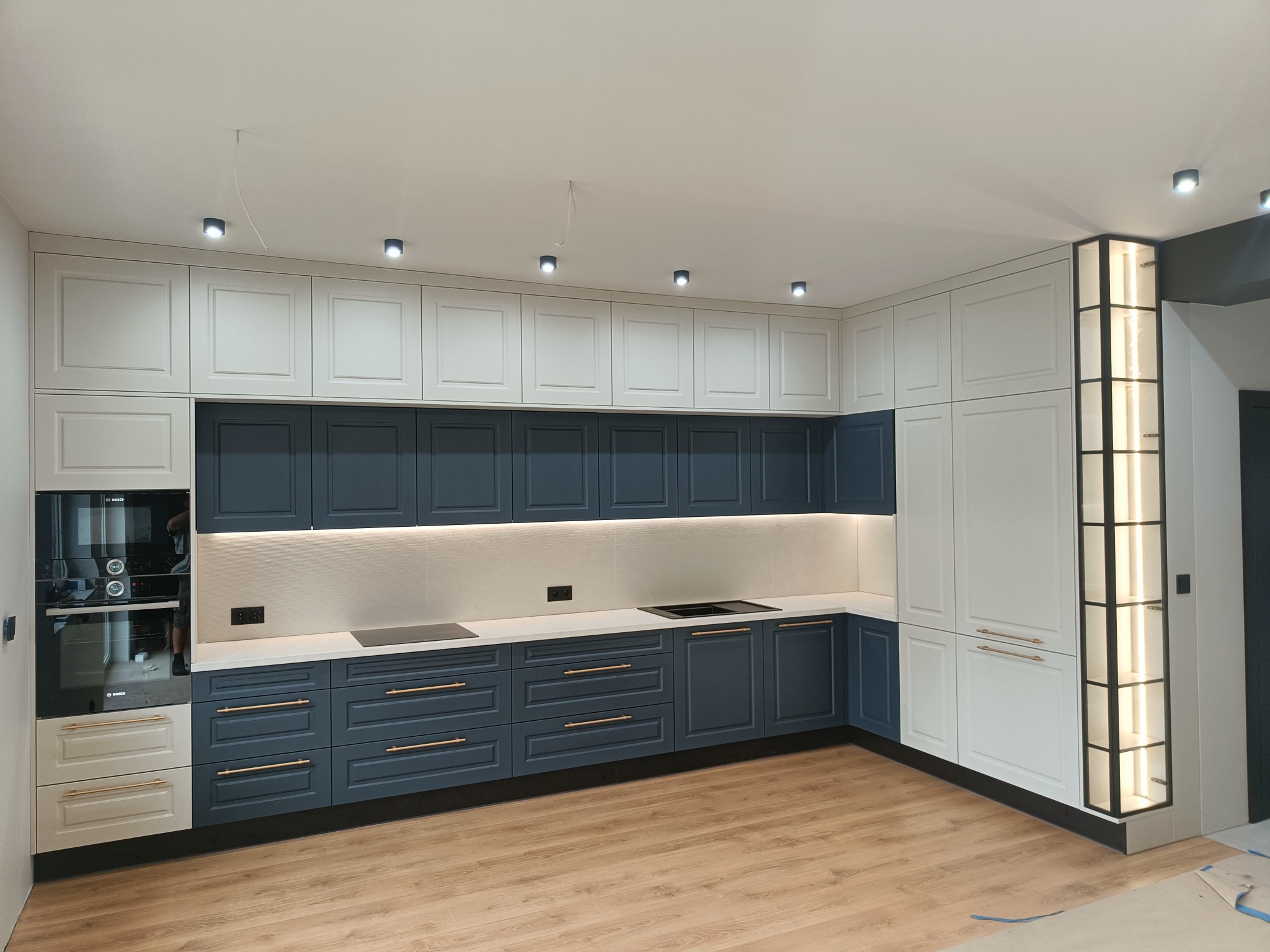 Nowoczesna kuchnia z granatowymi dolnymi szafkami i białymi górnymi, oświetleniem LED pod szafkami i drewnianą podłogą. Widoczny piekarnik i płyta indukcyjna.