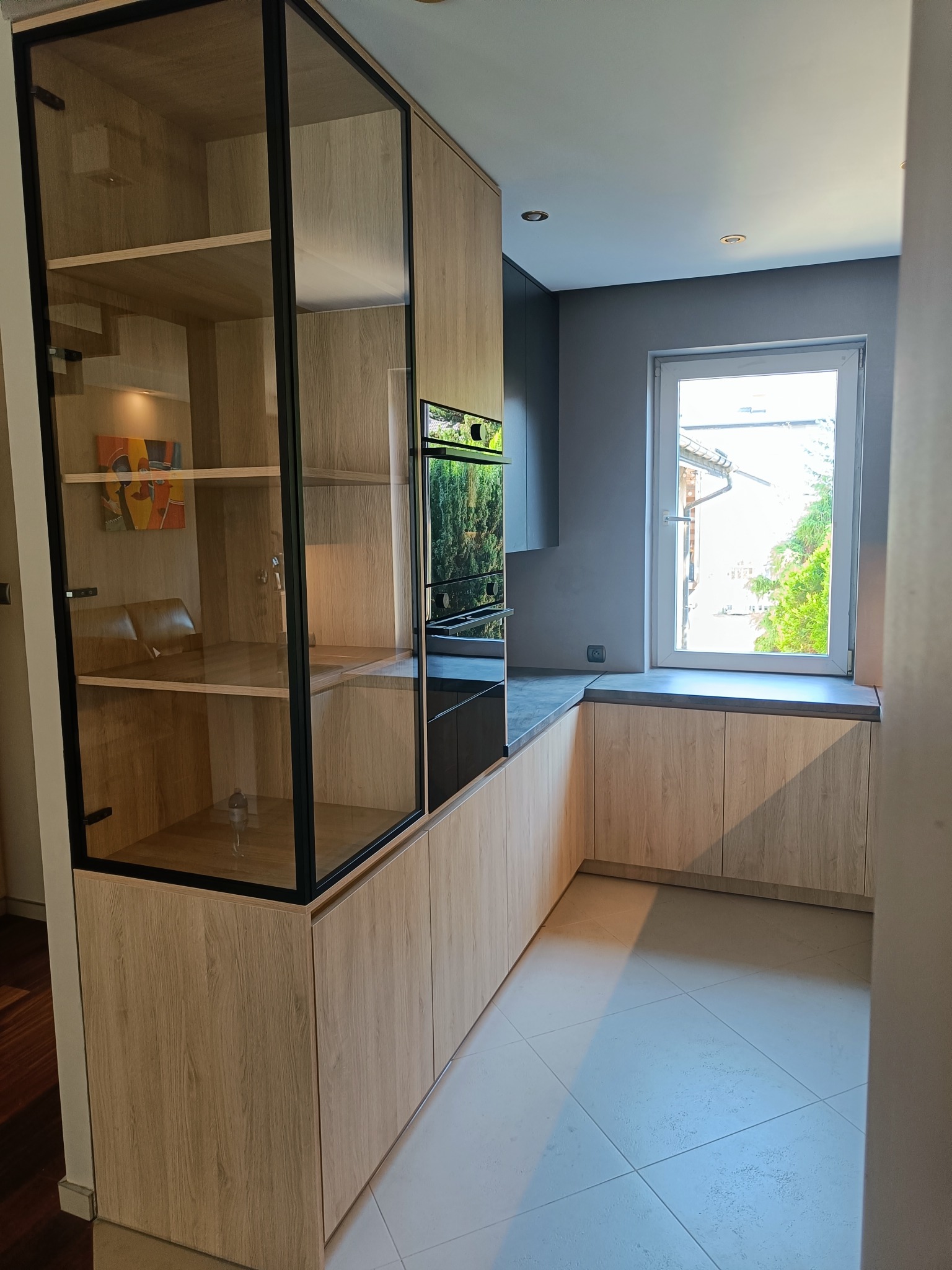 Jasna kuchnia z drewnianymi szafkami i czarnymi akcentami, przeszklona witryna z obrazem w środku, szare ściany i okno z widokiem na zieleń.