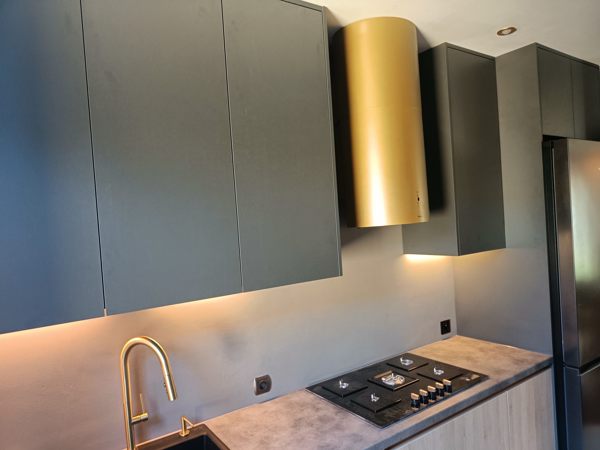 Nowoczesna kuchnia z szarymi szafkami, złotym okapem i baterią. Podświetlenie LED podkreśla minimalistyczny design. Widoczna płyta grzewcza i fragment lodówki.