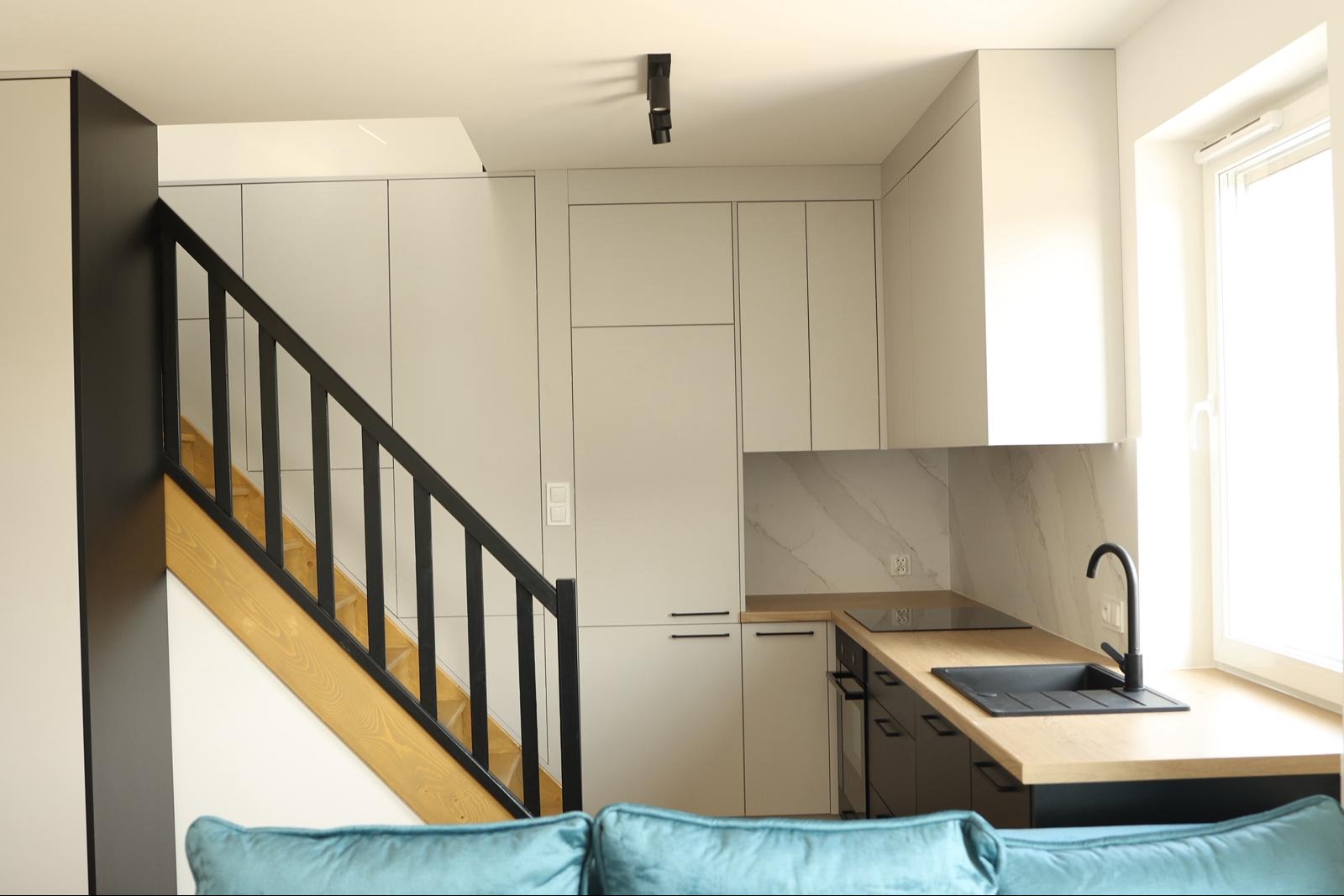 Jasna kuchnia z zabudową pod schodami, czarne akcenty: bateria, relingi, schody. Blat drewniany, marmurowy backsplash. Widok z perspektywy sofy.