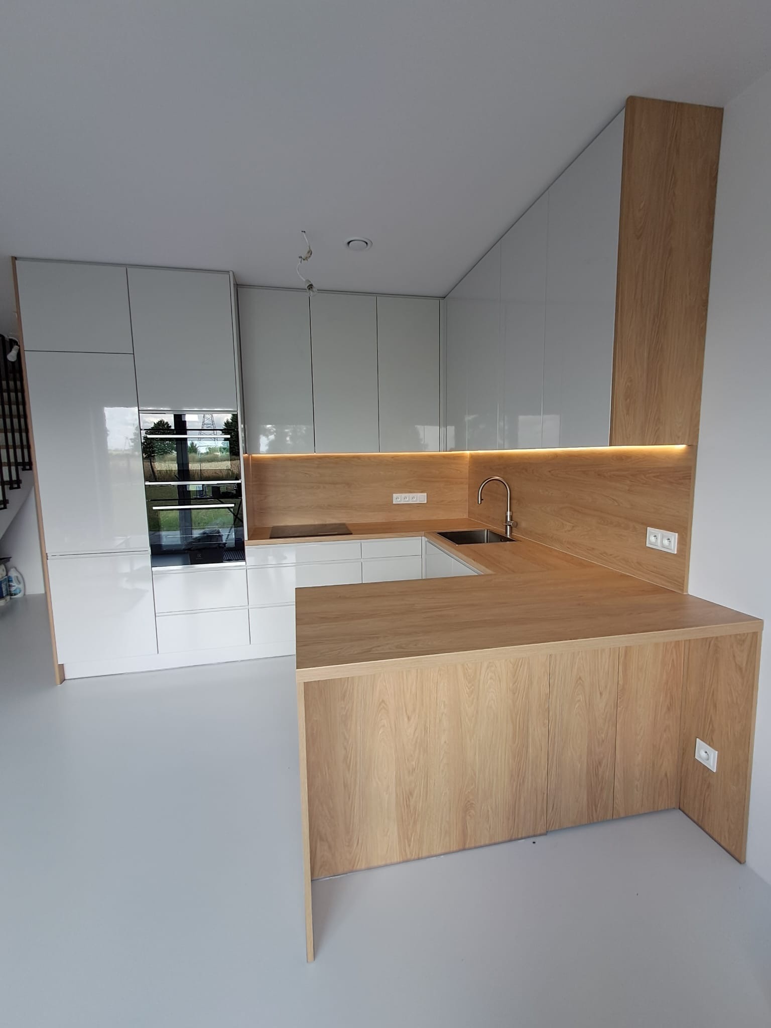 Nowoczesna kuchnia z białymi, lakierowanymi szafkami i drewnianym blatem z oświetleniem LED. Widoczny zlew i bateria. Minimalistyczny design.