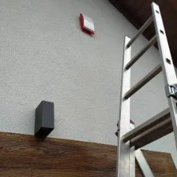 Zewnętrzna ściana domu z zamontowaną sygnalizacją alarmową z czerwoną obudową, czarną lampą oraz drabina oparta o ścianę.