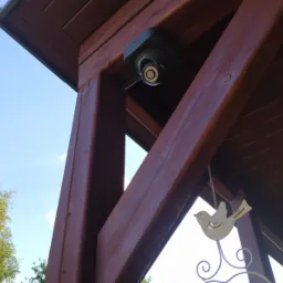 Kamera monitoringu kopułkowa zamontowana pod drewnianą konstrukcją altany, widok z dołu, dekoracja w kształcie ptaka.