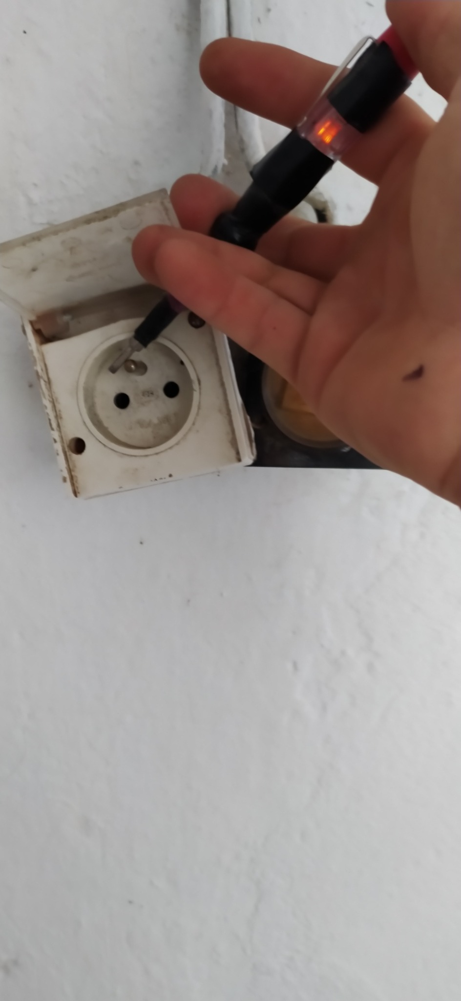 Elektryk sprawdza napięcie w brudnym gniazdku elektrycznym za pomocą próbnika napięcia, którego dioda LED świeci na pomarańczowo.