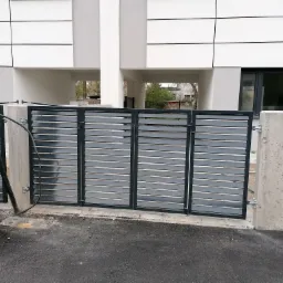 Nowoczesna, pozioma brama wjazdowa z szarych profili, zamontowana na betonowych słupkach, widoczna na tle budynku z białymi panelami.