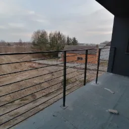 Czarna, pozioma balustrada z cienkich prętów na balkonie nowego budynku, widok na teren budowy i ceglany budynek w tle.