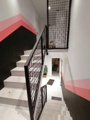 Nowoczesne schody wewnętrzne z metalową balustradą o geometrycznym wzorze, malowane ściany w pasy czerni i różu, widok z góry schodów w dół.