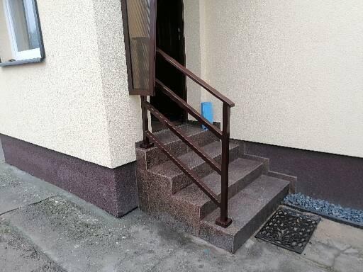 Zewnętrzne schody wejściowe z brązową metalową barierką, wykończone płytkami imitującymi kamień, prowadzące do drzwi wejściowych, z wycieraczką przed drzwiami.