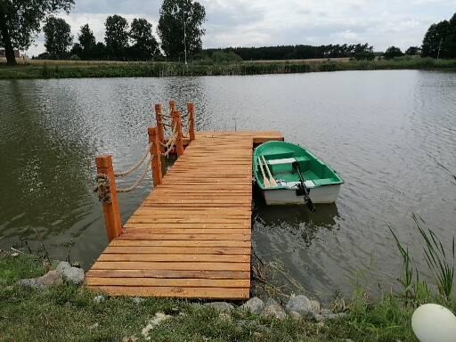 Drewniany pomost z linami jutowymi jako barierką, prowadzący do jeziora z zacumowaną zieloną łódką.