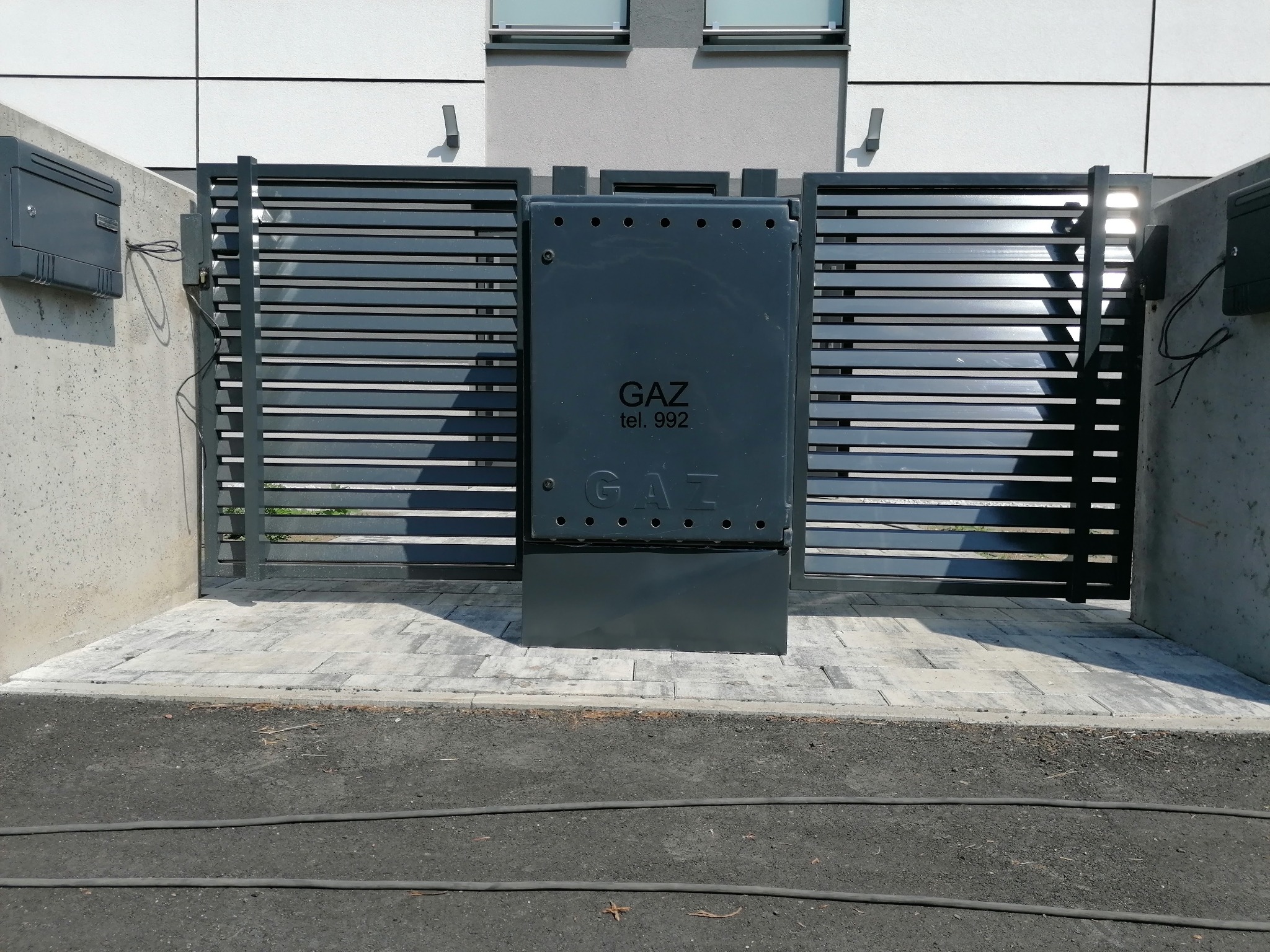 Nowoczesna, pozioma brama wjazdowa w kolorze grafitowym, z widoczną skrzynką gazową pośrodku, zamontowana przed budynkiem o jasnej elewacji.