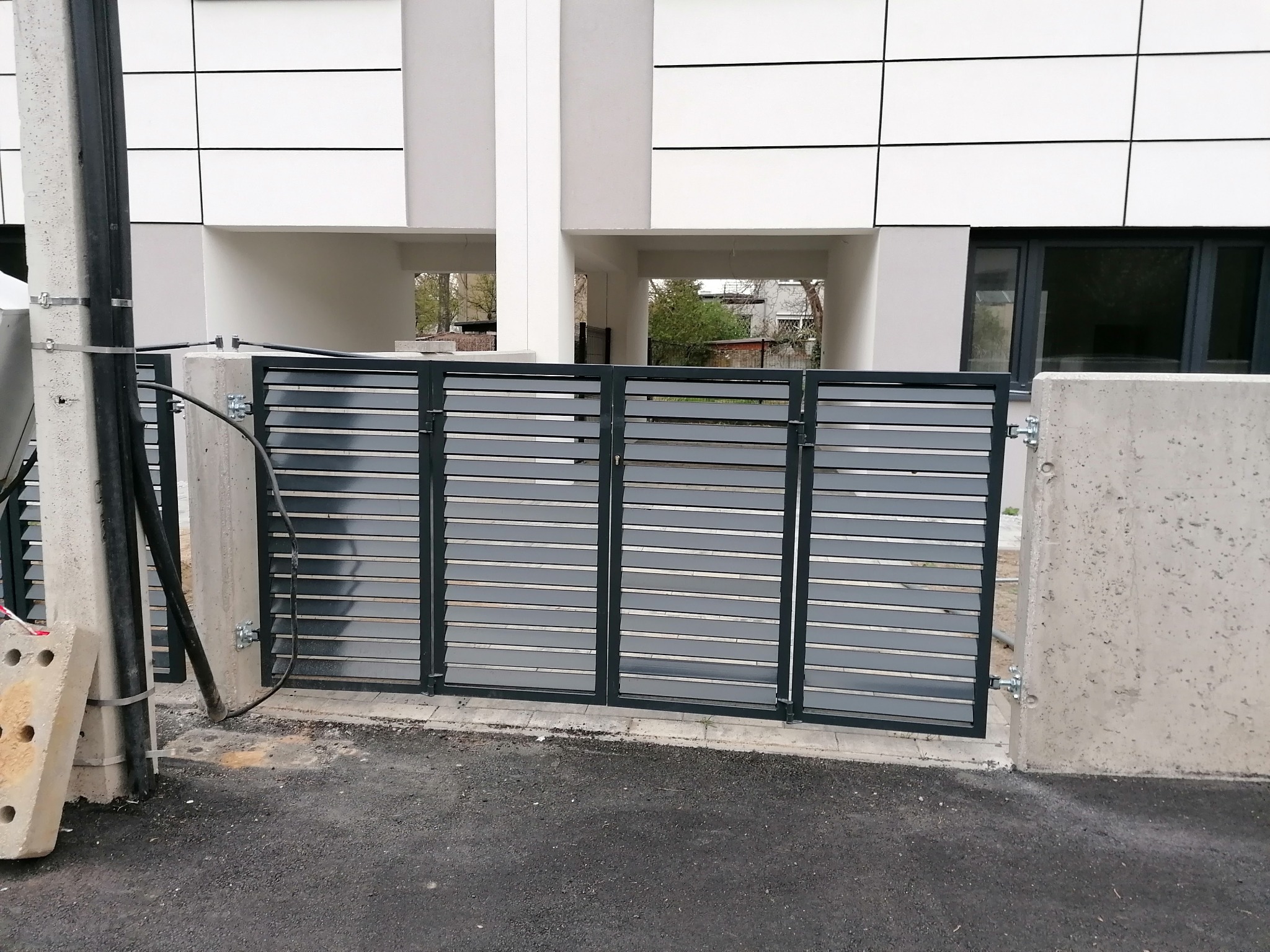 Nowoczesna, pozioma brama wjazdowa z szarych profili, zamontowana na betonowych słupkach, widoczna na tle budynku z białymi panelami.