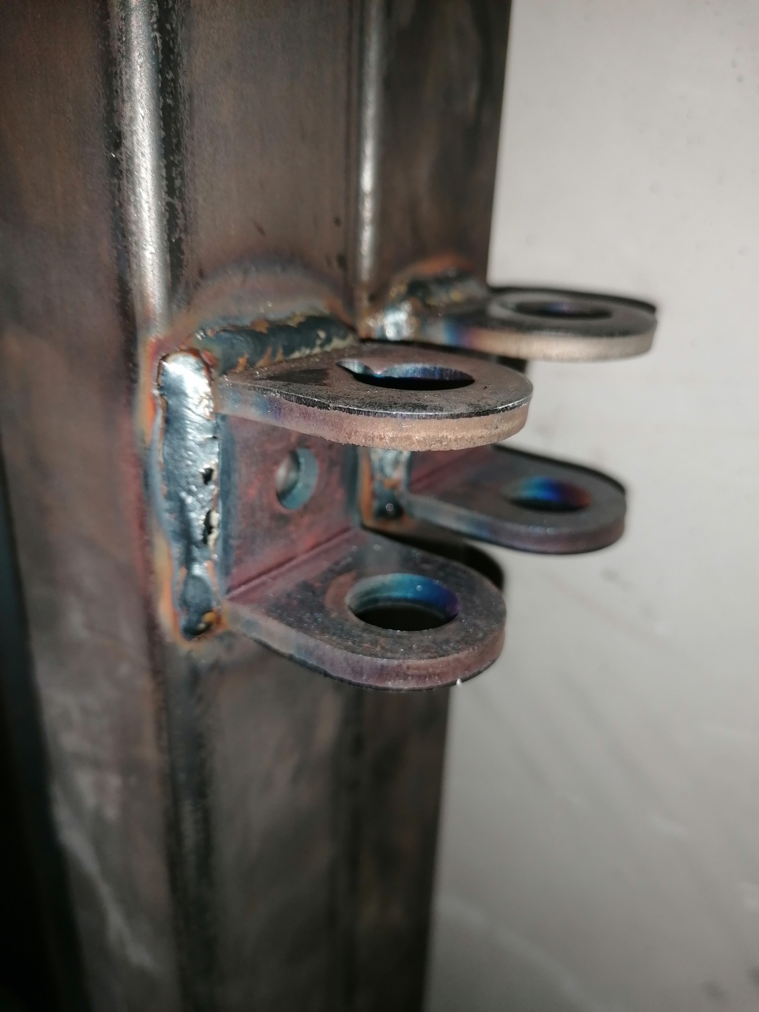 Metalowy słupek z przyspawanymi trzema uchwytami z otworami, widoczne ślady spawania i przebarwienia termiczne na metalu.