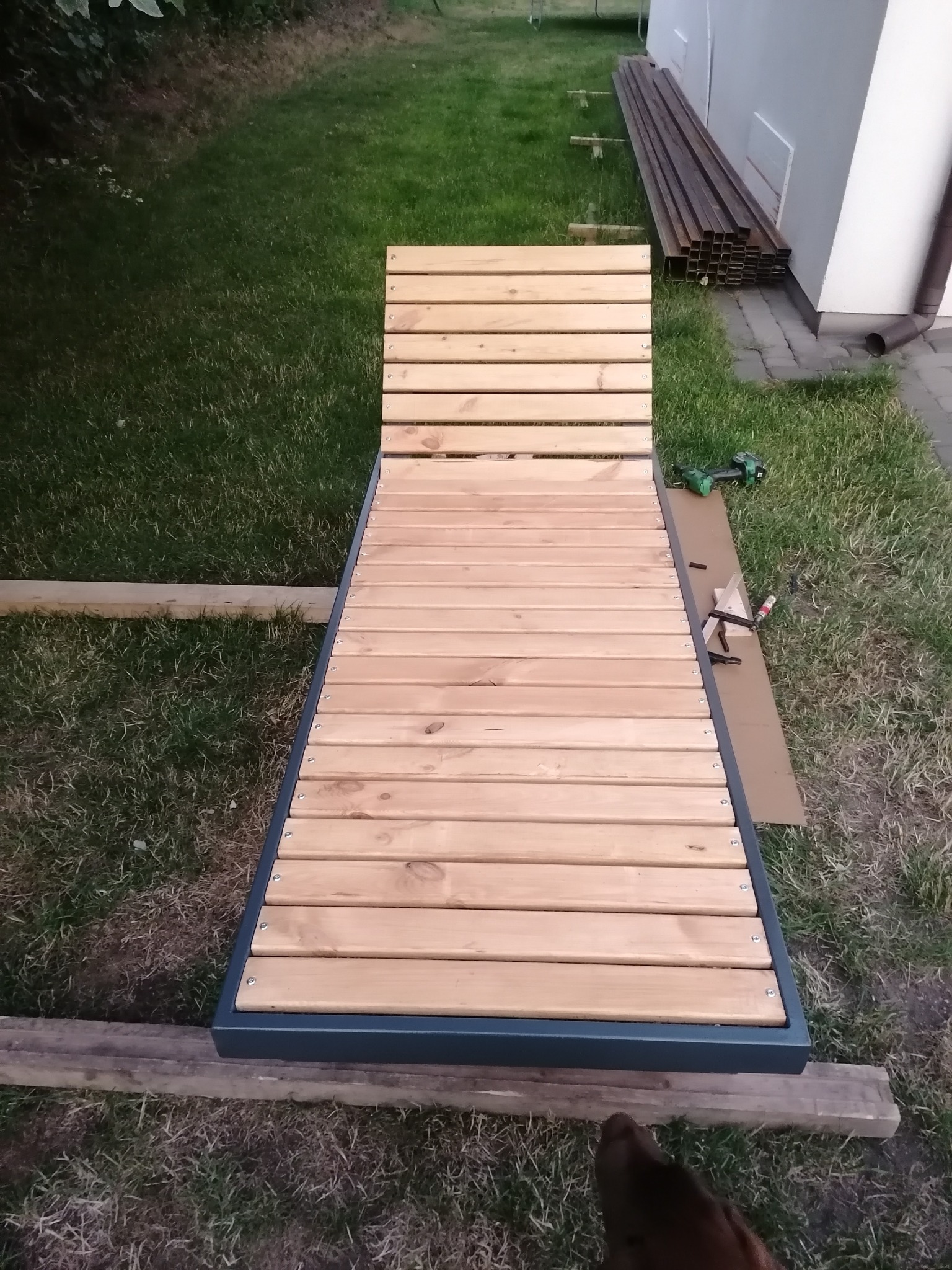 Konstrukcja leżaka ogrodowego z drewnianych listew i metalowej ramy, w trakcie montażu na trawniku, widoczne narzędzia i elementy konstrukcyjne.