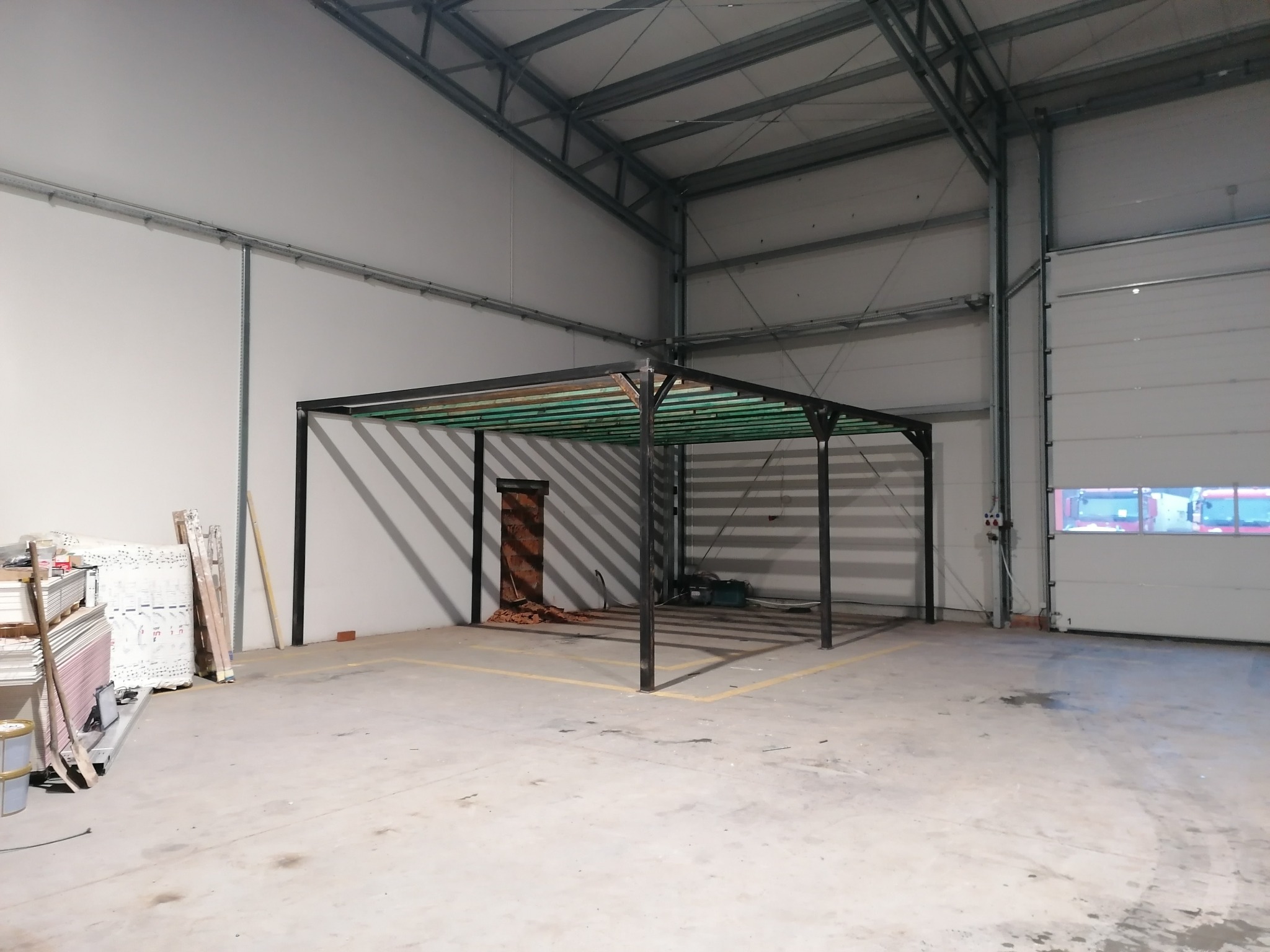 Metalowa konstrukcja z drewnianymi elementami w hali produkcyjnej, z widocznymi materiałami budowlanymi i żółtymi liniami na podłodze, wskazującymi obszar roboczy.