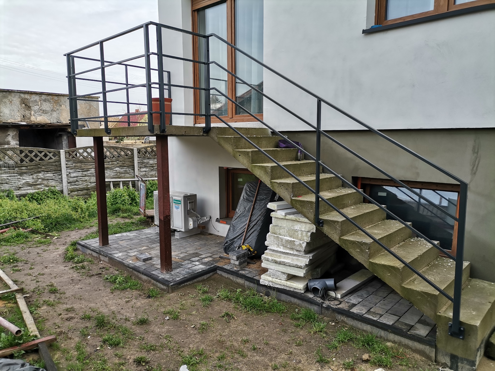Betonowe schody zewnętrzne z metalową balustradą prowadzące do drzwi na podwyższeniu; widoczne materiały budowlane i kostka brukowa w trakcie układania.