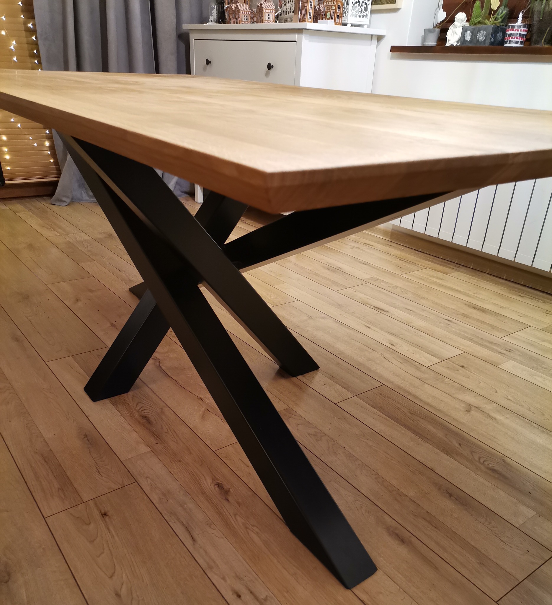 Stół z blatem z jasnego drewna i czarnymi, metalowymi nogami w kształcie litery X, na tle drewnianej podłogi i mebli w stylu skandynawskim.