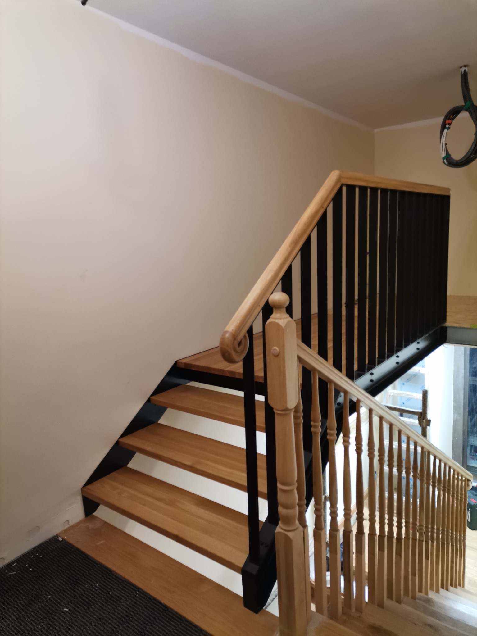 Drewniane schody z czarną metalową konstrukcją i jasną balustradą, widok z dołu, na ścianie widoczne przewody elektryczne.