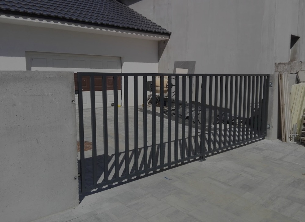 Nowoczesna, metalowa brama wjazdowa z pionowymi prętami, zamontowana na betonowych słupkach przy posesji z garażem i szarą kostką brukową.