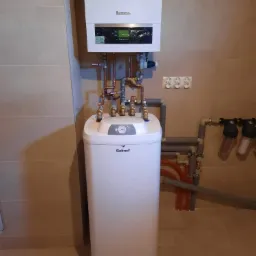 Kompaktowy zestaw: wiszący kocioł grzewczy Buderus nad stojącym podgrzewaczem wody Galmet, z widocznymi rurami miedzianymi i filtrami wody na tle beżowych płytek.