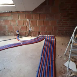 Montaż instalacji ogrzewania podłogowego w surowym stanie: widoczne rury w kolorze niebieskim i czerwonym, prowadzone wzdłuż ceglanej ściany poddasza, drabina z wiertarką w tle.