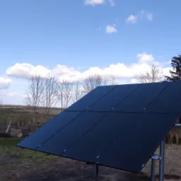 Panel słoneczny ustawiony na metalowych wspornikach na tle błękitnego nieba z chmurami, widoczne drzewa i budynek w oddali.