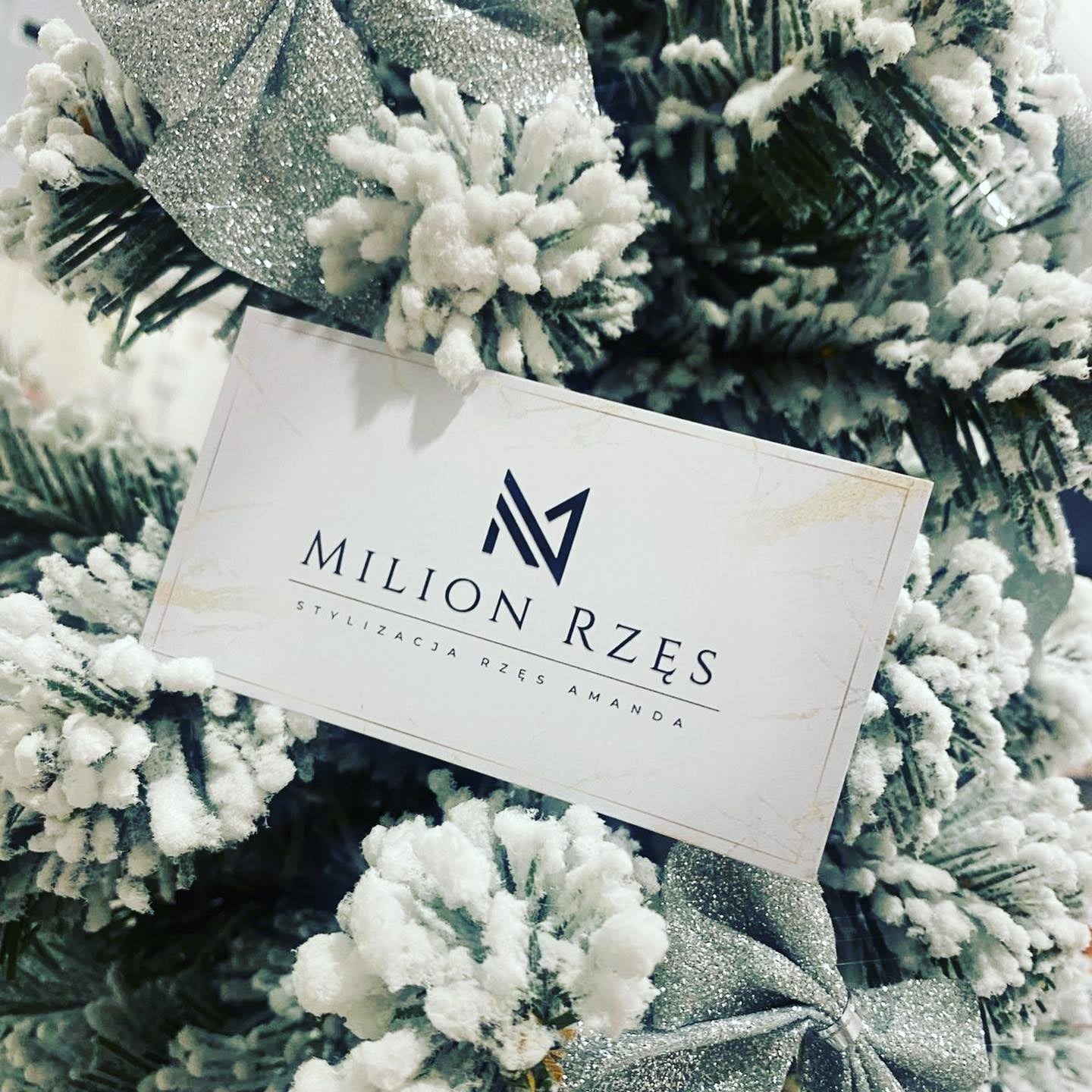 Wizytówka salonu stylizacji rzęs 'Milion Rzęs Amanda' umieszczona na tle sztucznego, ośnieżonego drzewka świątecznego z srebrnymi kokardami.