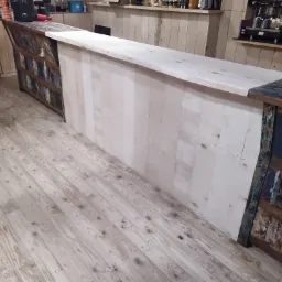 Bar wykonany z jasnego drewna z elementami dekoracyjnymi w stylu rustykalnym, widoczny dzwonek, drewniana podłoga i ekspres do kawy w tle.